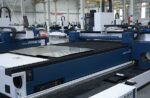 A דגם C Series blue and white industrial laser cutting machine in a spacious factory, with metal sheets on its bed; the setting features high ceilings, white walls, and visible support beams.
