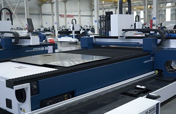 A דגם C Series blue and white industrial laser cutting machine in a spacious factory, with metal sheets on its bed; the setting features high ceilings, white walls, and visible support beams.