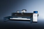 The דגם 24030TX is a SENFENG industrial laser cutting machine featuring a large flat cutting bed and a right-side control panel, shown against a gradient blue background.