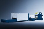 The דגם 1502CH is a large industrial laser cutting machine with a sleek white and blue exterior, featuring a long flatbed, control units, and an attached roll feeder, ideal for clean, well-lit industrial settings.