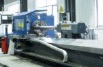 The דגם LYCK8463 industrial CNC lathe operates in a factory, partially machining a long metal workpiece as metal shavings gather on the bed. Safety warning signs are clearly visible on the machine.