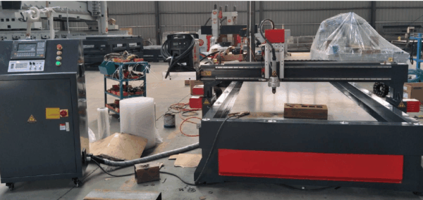 A CNC plasma cutting machine with a control panel on the left, metal cutting bed in the center, and various tools and materials scattered around in an industrial workshop setting.