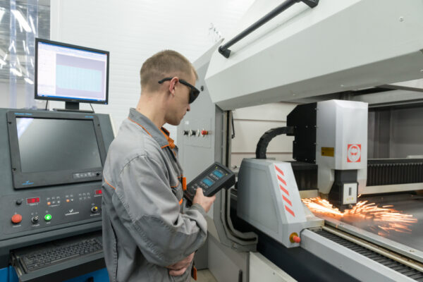 A person wearing safety glasses operates a control device next to a laser cutting machine, which is emitting sparks as it cuts metal. Computer screens and industrial equipment are visible in the background.