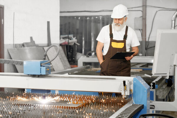 An older man in a white hard hat and overalls stands in a factory, holding a tablet and observing a CNC machine cutting metal with bright sparks flying from the cutting area.