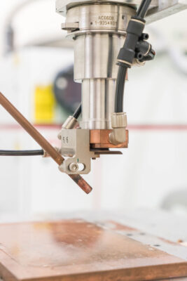 A close-up of an industrial robotic arm performing a welding or soldering operation on a metal workpiece in a manufacturing environment.