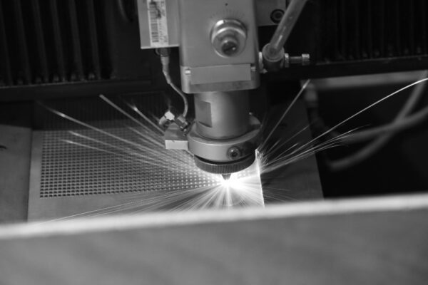 A close-up view of a laser cutting machine emitting bright sparks as it cuts through a sheet of metal, with a grid pattern visible on the metal surface.