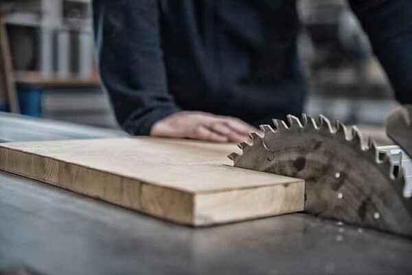 Person wearing dark clothing guiding a piece of wood towards a circular saw blade on a workbench, preparing to cut the wood in a woodworking shop.