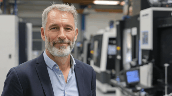 A middle-aged man with gray hair and a beard, wearing a blue suit and light blue shirt, stands smiling in a modern industrial or manufacturing facility with machines in the background.